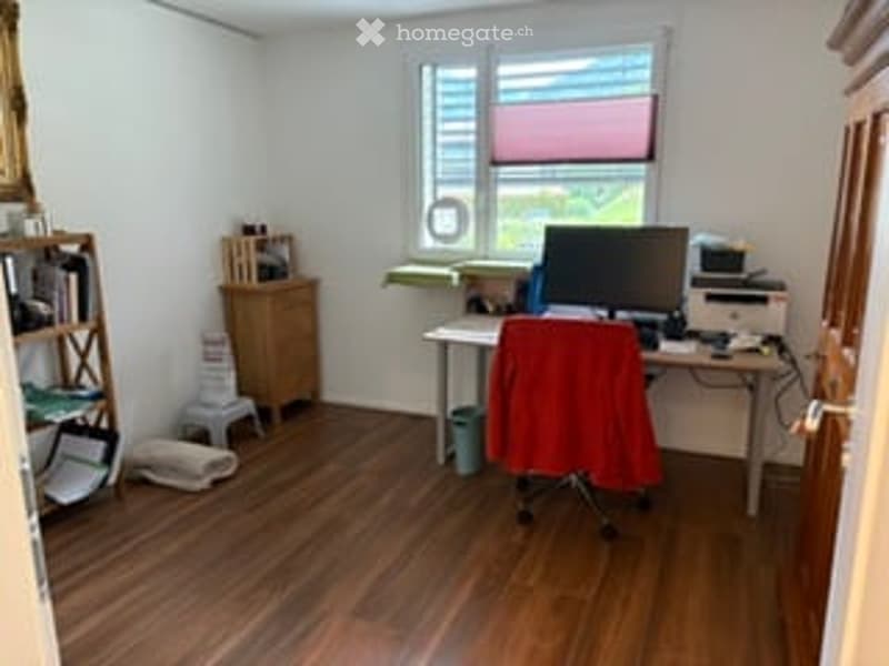 A small, well-lit room with wooden flooring, a desk with a computer monitor, a red chair, a printer, a bookshelf, and a small cabinet.