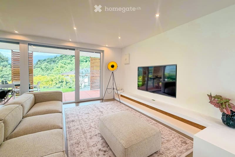 Modern living room with large sliding glass doors offering a scenic view, a grey sectional sofa, ottoman, flat-screen TV, floor lamp, and a small table with a plant.