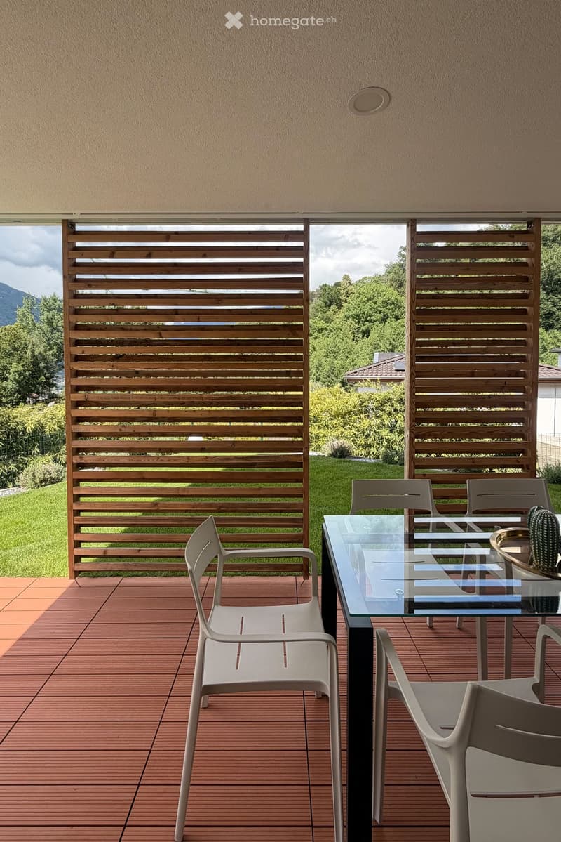 Balcony with wooden slatted screens, glass table, and white plastic chairs, overlooking a green garden and mountains.