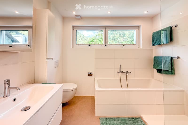 Modern bathroom with a white sink, toilet, bathtub, and large windows offering a scenic view. Green towels are neatly hung on a rack, and the space is well-lit with recessed ceiling lights.