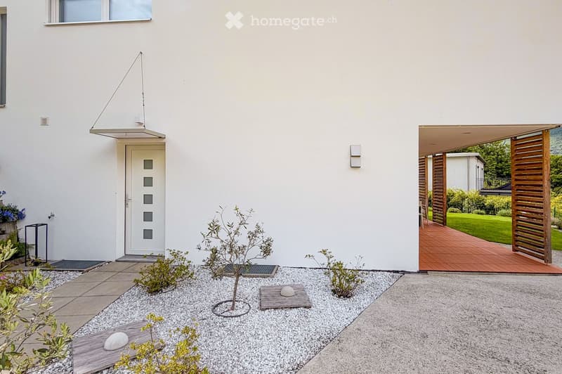 Modern garden area with white house facade, small tree, gravel, wooden deck, and covered patio area.