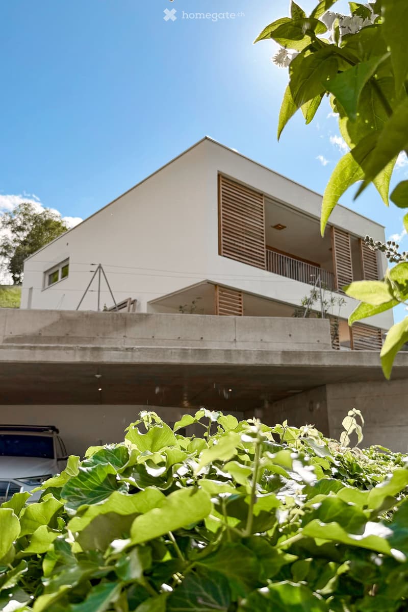 Modern two-story house with white exterior, large windows, and a balcony on the upper level. The house features a flat roof and is surrounded by greenery.