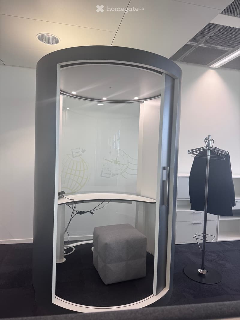 A modern, enclosed office space with a round, glass-walled booth. Inside, there is a grey stool, a sleek white table with cables, and a coat rack with a dark jacket hanging on it. The booth features a contemporary design with a black and white color schem