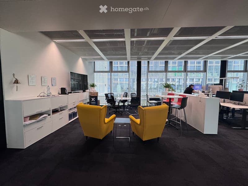 Modern office space with large windows, yellow chairs, a small table, desks, and computer setups.