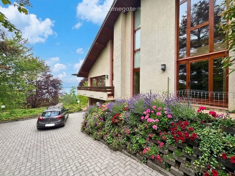 Two-story house with a sloped roof, large windows, and a balcony. The front yard features a well-maintained garden with colorful flowers and a paved driveway with a parked car.