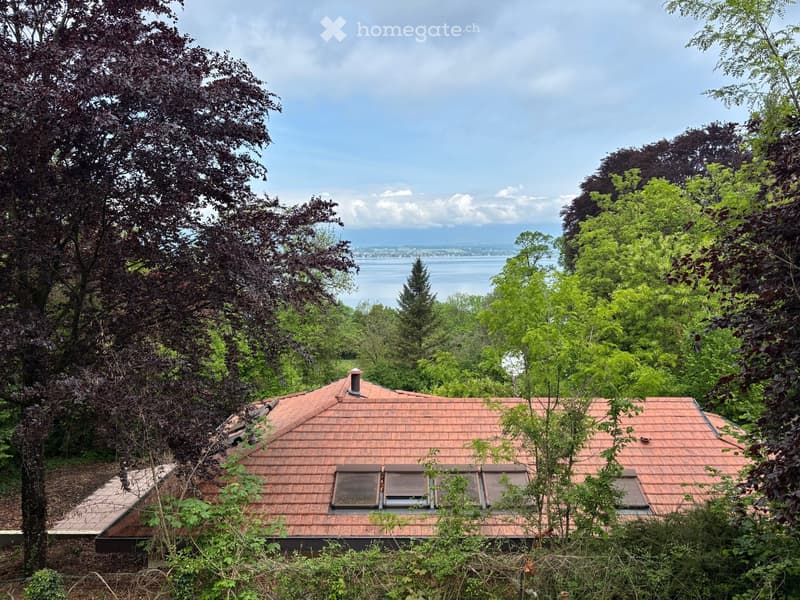 A two-story house with a red-tiled roof, surrounded by lush greenery and trees, overlooking a body of water.