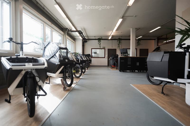 Bright, modern showroom with bicycles and electric cargo bikes displayed, large windows, wooden flooring, and a cabinet with a screen and chair.