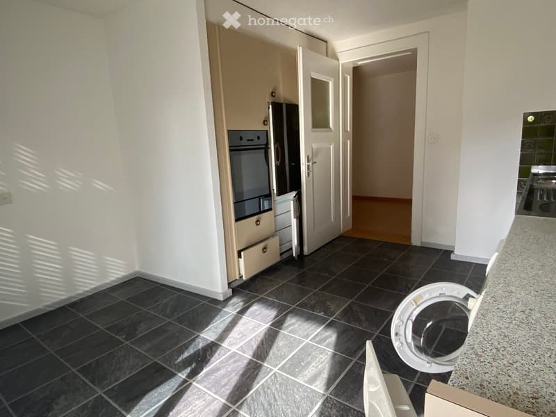 Modern kitchen with dark tiled flooring, white walls, and built-in appliances including a refrigerator, oven, and washing machine. Cabinets are light-colored, and the space is well-lit with natural light.