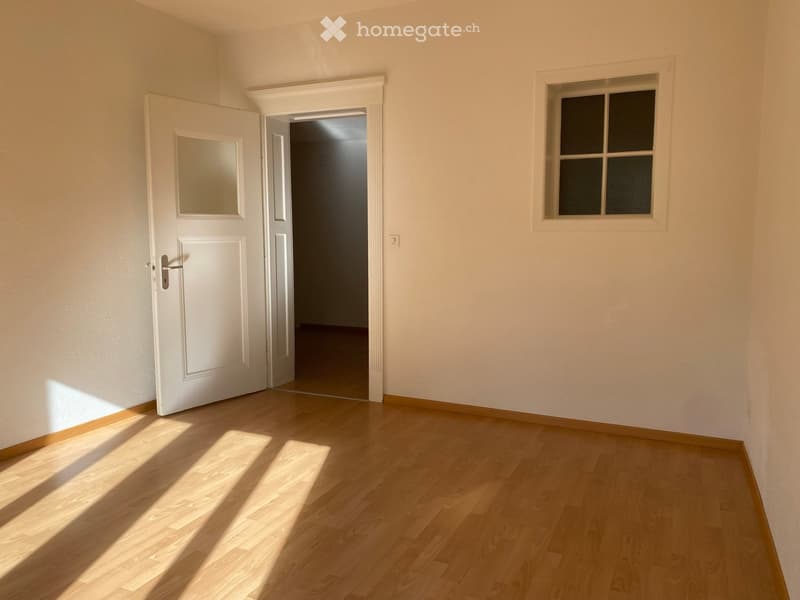 Empty room with light wooden flooring, white walls, a white door with a mirror panel, and a window with four panes.