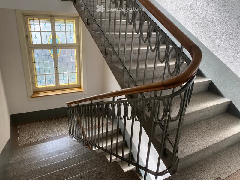 Spiral staircase with metal railing and wooden handrail, leading to a window with stained glass.