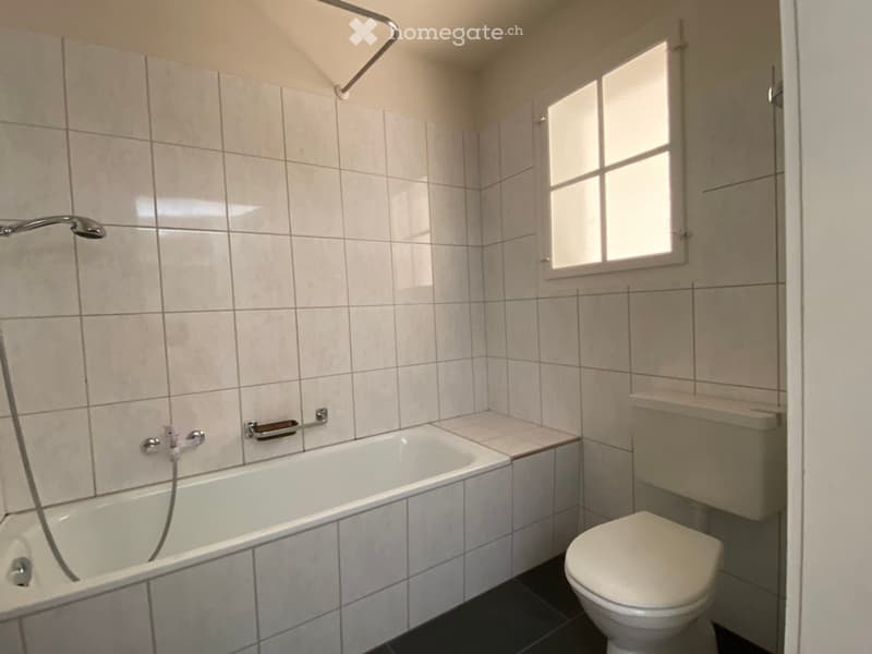 Bathroom with a bathtub, toilet, and a small window. The walls are tiled, and there is a handrail next to the bathtub.