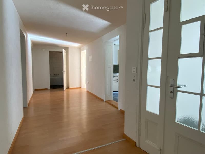 Empty hallway with wooden flooring, white walls, and recessed lighting. Doors lead to adjacent rooms, including a kitchen area visible in the background.
