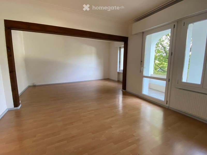 Empty room with wooden flooring, white walls, large windows, and a door leading to another space.