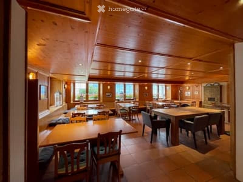 Restaurant or dining area with wooden tables, chairs, and benches arranged around the space. The room features exposed wooden beams on the ceiling and large windows letting in natural light.