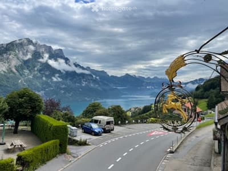 Scenic outdoor area with a road leading to a parking lot, surrounded by lush greenery and a stunning mountain lake view.