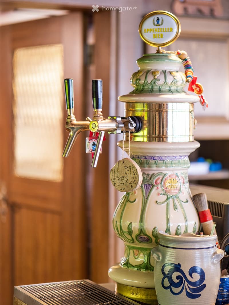 A beer tap with a decorative ceramic jug and mug, featuring a sign reading "Appenzeller Bier" and a small hanging ornament.