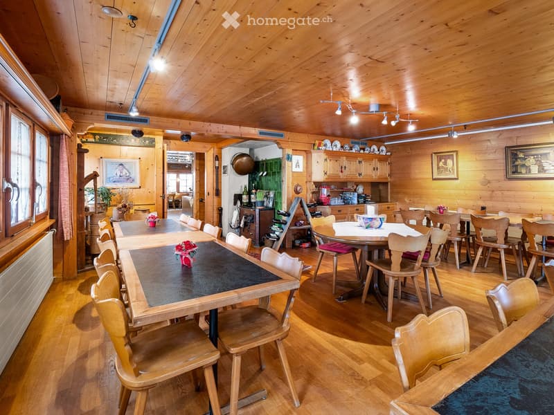 A spacious, wooden-paneled dining area with multiple tables and chairs, a kitchen area with cabinets and appliances, and decorative elements like framed pictures and flower vases.