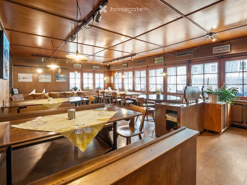 Restaurant interior with wooden tables, chairs, and large windows. Wooden ceiling and walls, pendant lights, and a potted plant on a counter.