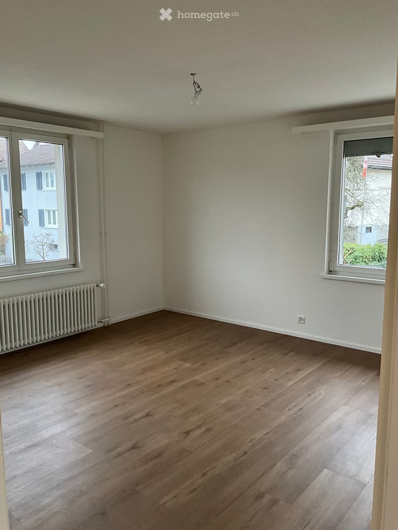 Empty room with light wooden flooring, white walls, and two windows with white frames. A radiator is visible under one window, and a ceiling light fixture is present.