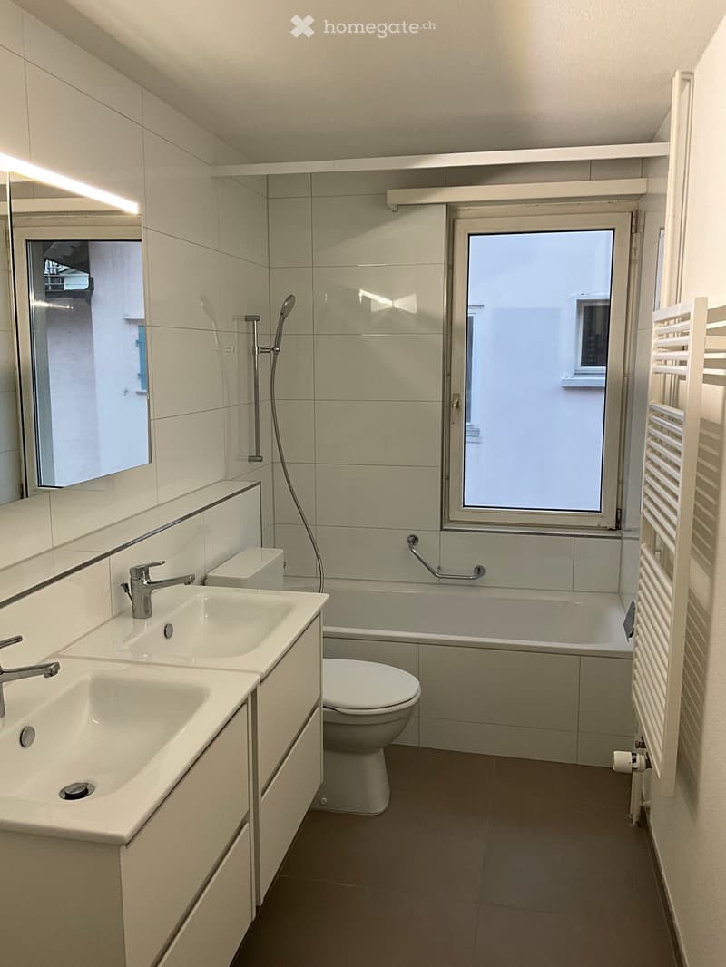 Modern bathroom with double sink, toilet, bathtub, and radiator. Features large windows and a sleek design.
