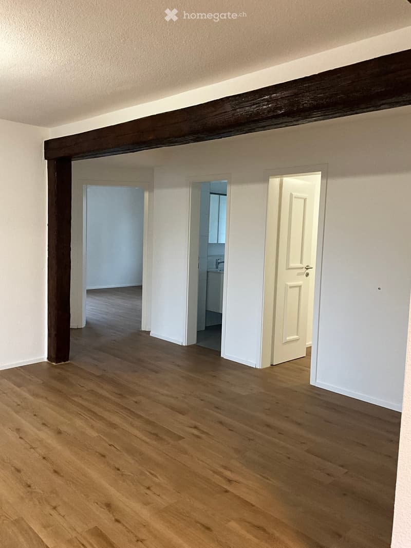 Empty room with wooden flooring, white walls, and exposed wooden beams. Three doors lead to other rooms, one of which shows a glimpse of a bathroom.