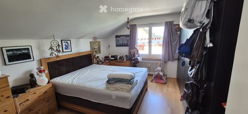 Cozy attic bedroom with wooden floors, slanted ceilings, and large windows. Features a double bed with a wooden headboard, nightstand, dreamcatchers, picture frames, and a small tent in the corner.
