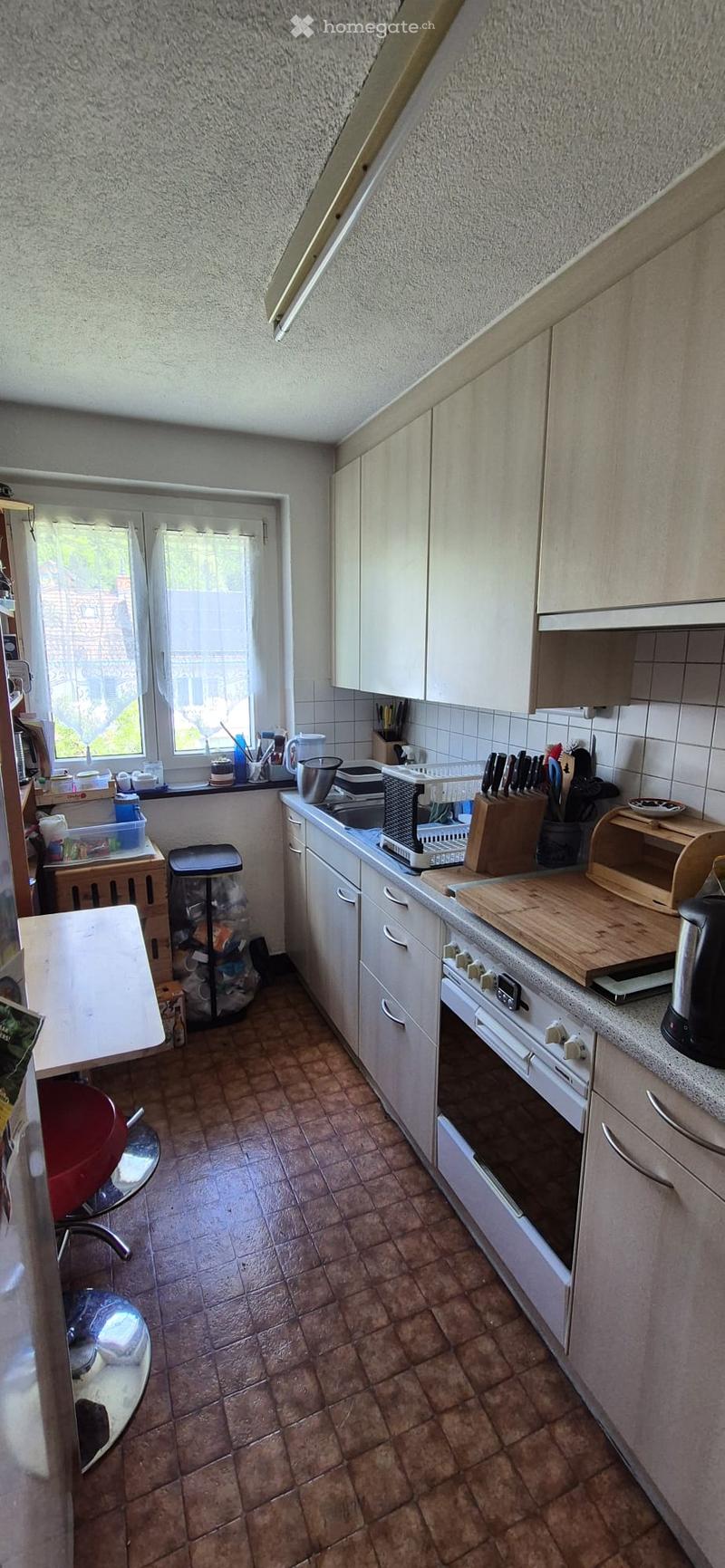 A small kitchen with white cabinets, a tiled floor, and various appliances including a stove, oven, and sink. There is a window with a view of the outside, a cutting board, and a knife block on the counter.