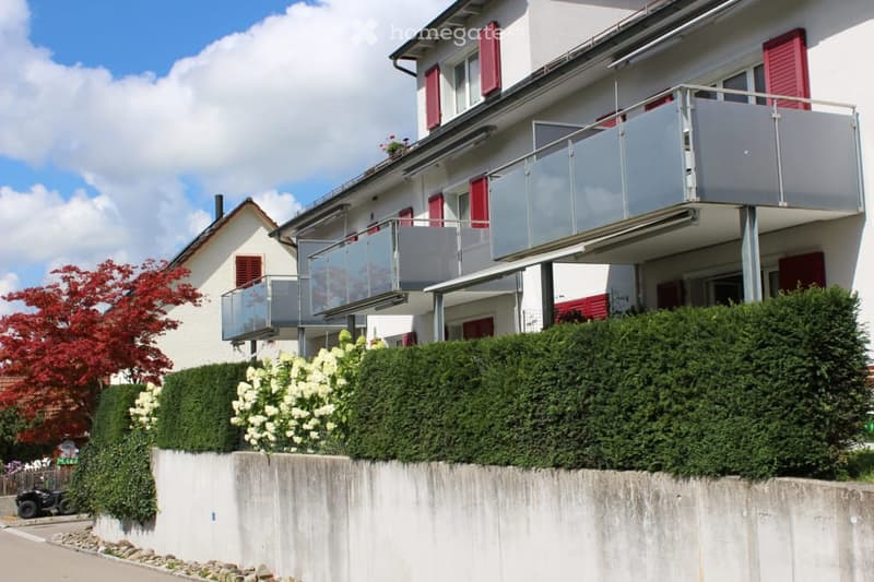 Two-story residential building with white walls, red shutters, and a large glass balcony overlooking a landscaped garden area.