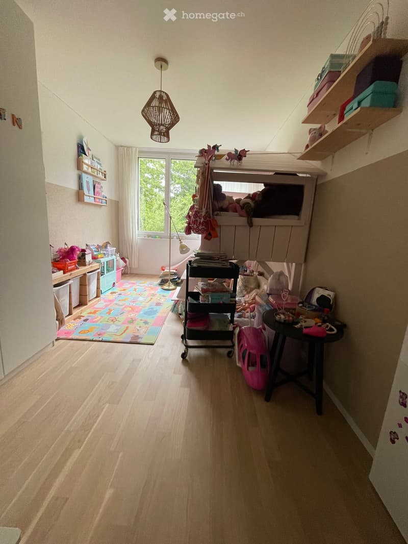 A well-lit children's room with wooden flooring, shelves, a small table, a chair, toys, and a colorful play mat.