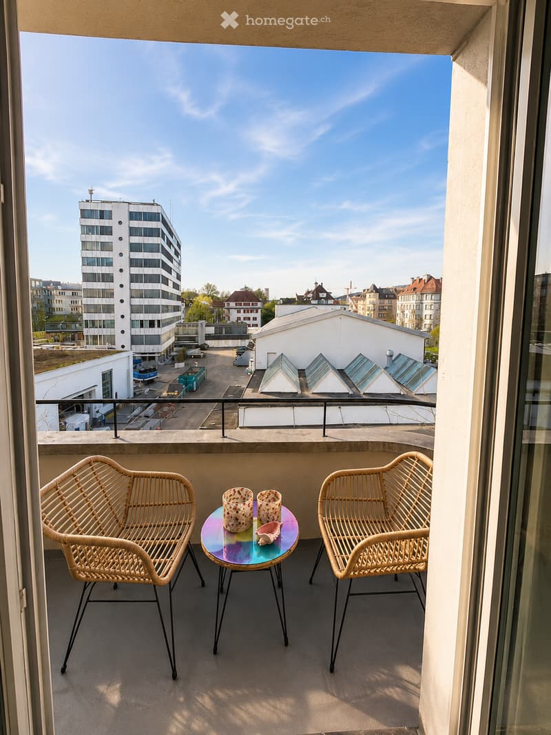 Balcony with two wicker chairs, a small round table with two cups and a seashell, overlooking a cityscape with modern and traditional buildings.