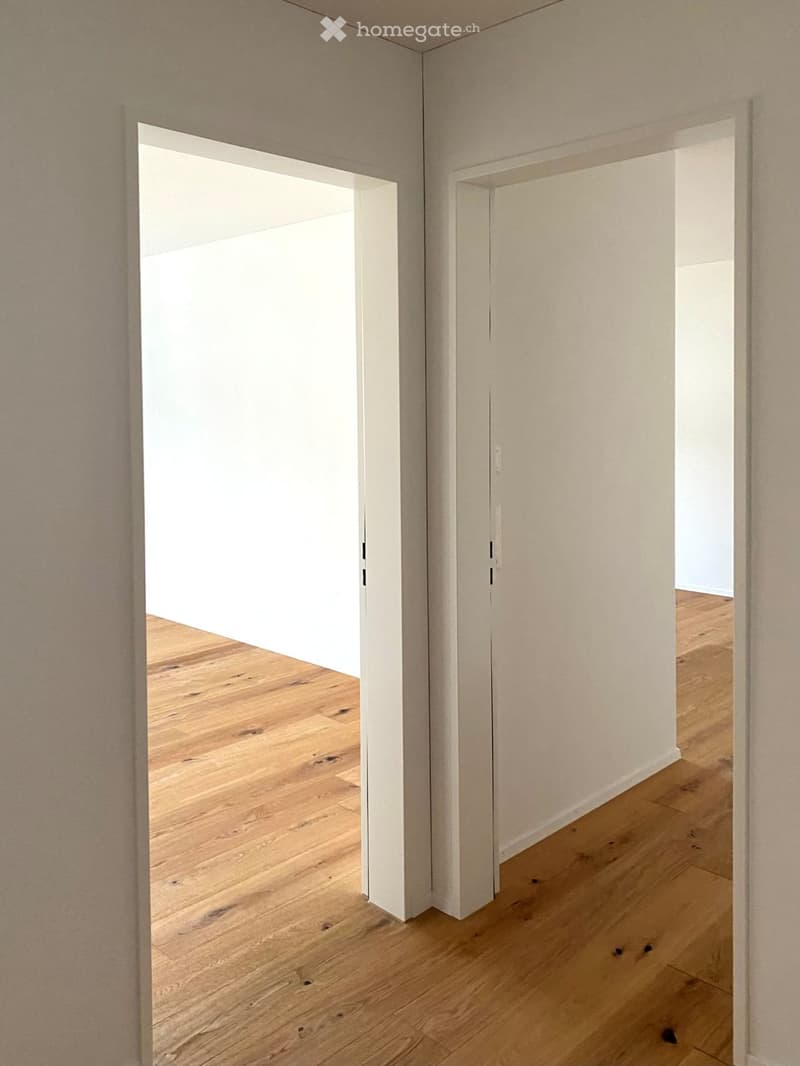 A modern, minimalist hallway with light wooden flooring and white walls, featuring an open doorway leading to another room.