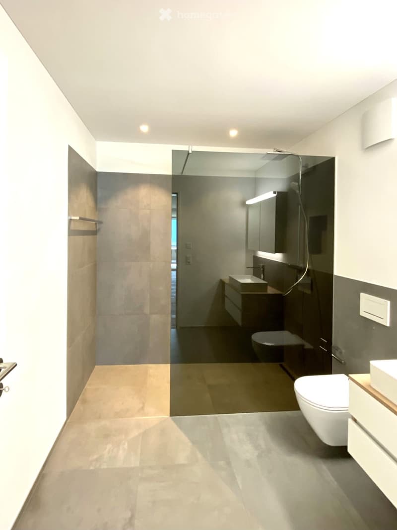 Modern bathroom with a large mirror, toilet, sink, and separate shower area with glass enclosure. Light-colored tiles and sleek fixtures.