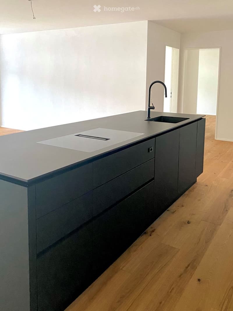 Modern kitchen with a dark grey countertop, built-in sink, and sleek faucet. Dark grey cabinets and light wooden flooring.