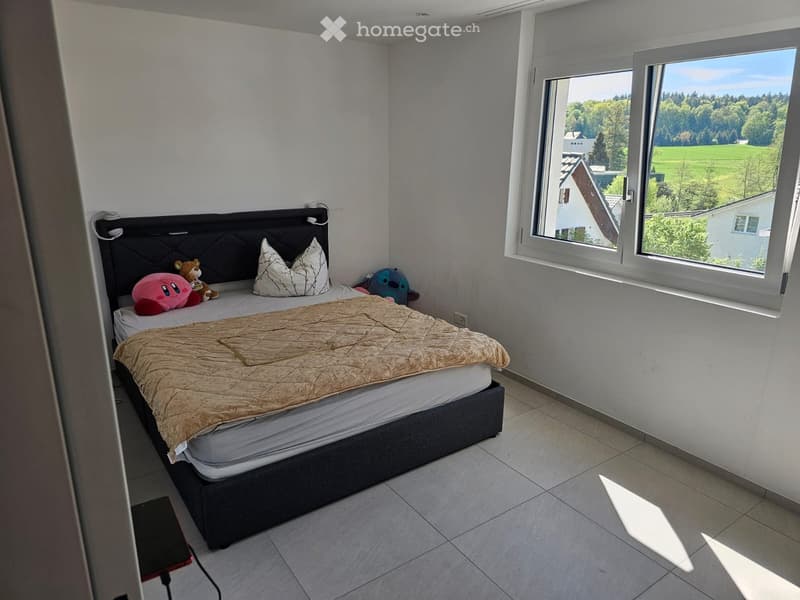 A modern bedroom with a large bed, headboard, pillows, and a beige blanket. The room features large windows with a scenic view of greenery and houses outside.