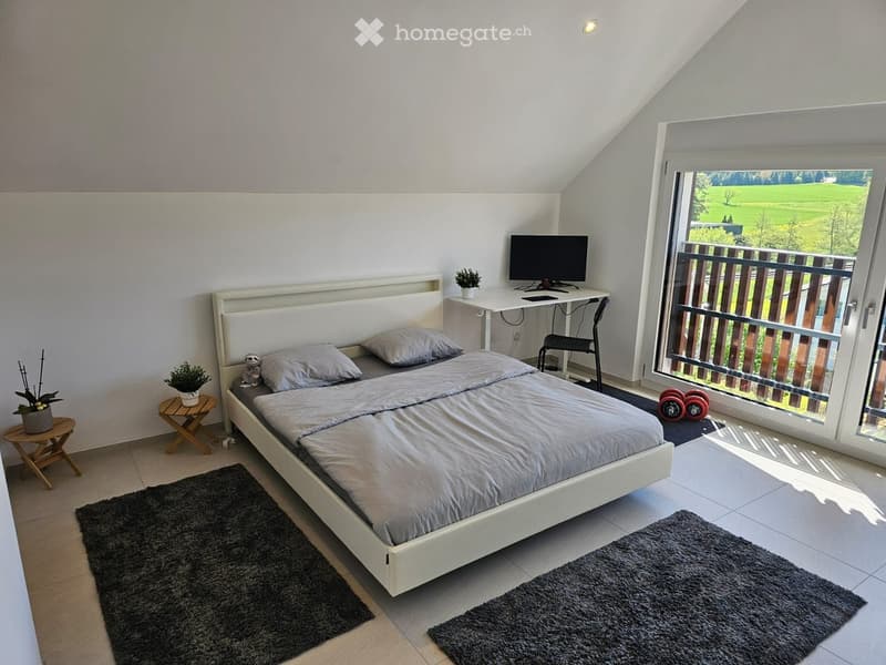 Modern bedroom with a white bed, grey bedding, a desk with a computer, a small stool, and a view of a green landscape through large windows.