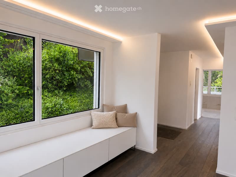 Modern, minimalist room with a built-in bench and decorative pillows, large windows overlooking greenery, sleek white walls, and wooden flooring.