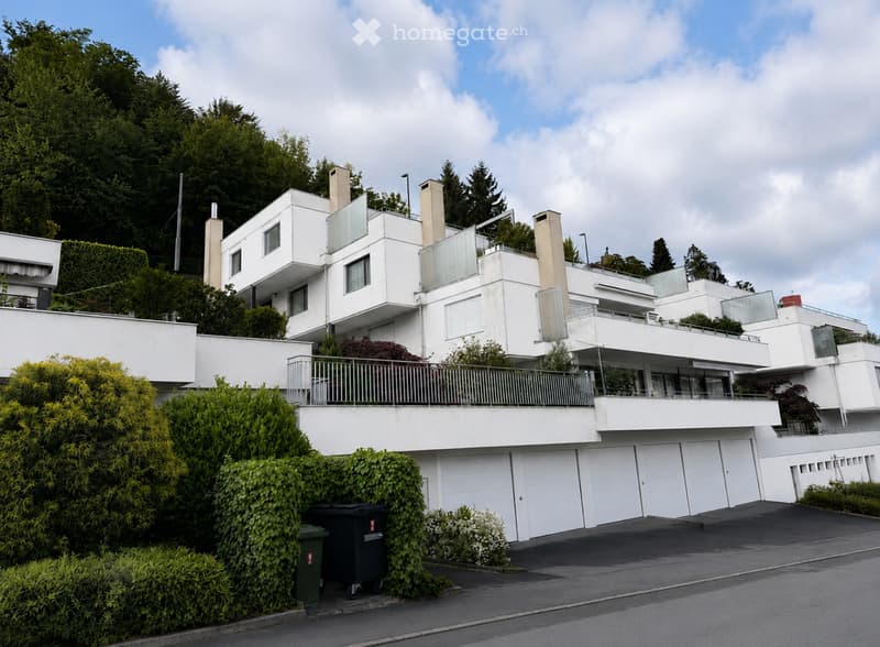 Modern white multi-level house with large windows, balconies, and a garage. Surrounded by greenery and trees.