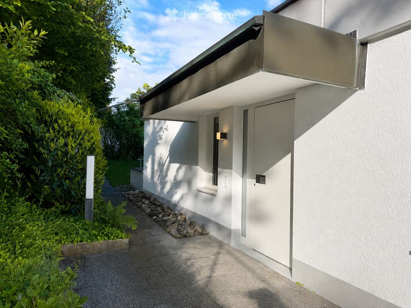 Modern white building with a sleek, minimalist design, featuring a covered entrance and a small garden area with greenery and a stone pathway.