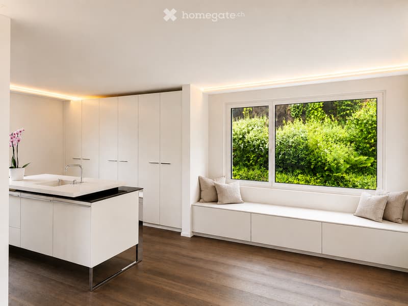 Modern kitchen with white cabinets, a built-in bench with pillows, and a large window overlooking greenery.