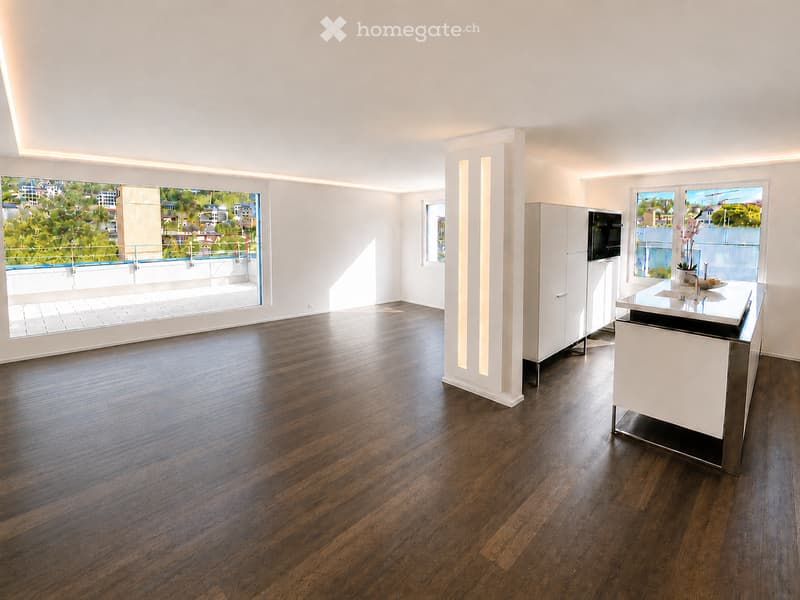Open-plan unfurnished room with wooden flooring, white walls, and large windows offering a view of a cityscape. Features include built-in cabinets, a modern kitchen island, and recessed lighting.