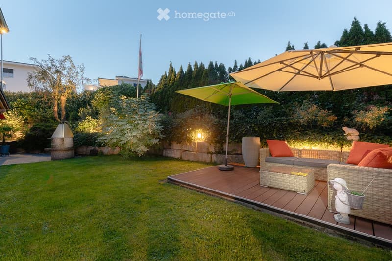 Well-maintained garden with a wooden deck, outdoor seating area, large umbrellas, and decorative lighting.