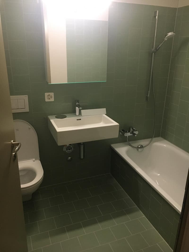 Modern bathroom with green tiled walls and floor, featuring a white sink, toilet, bathtub, and showerhead.