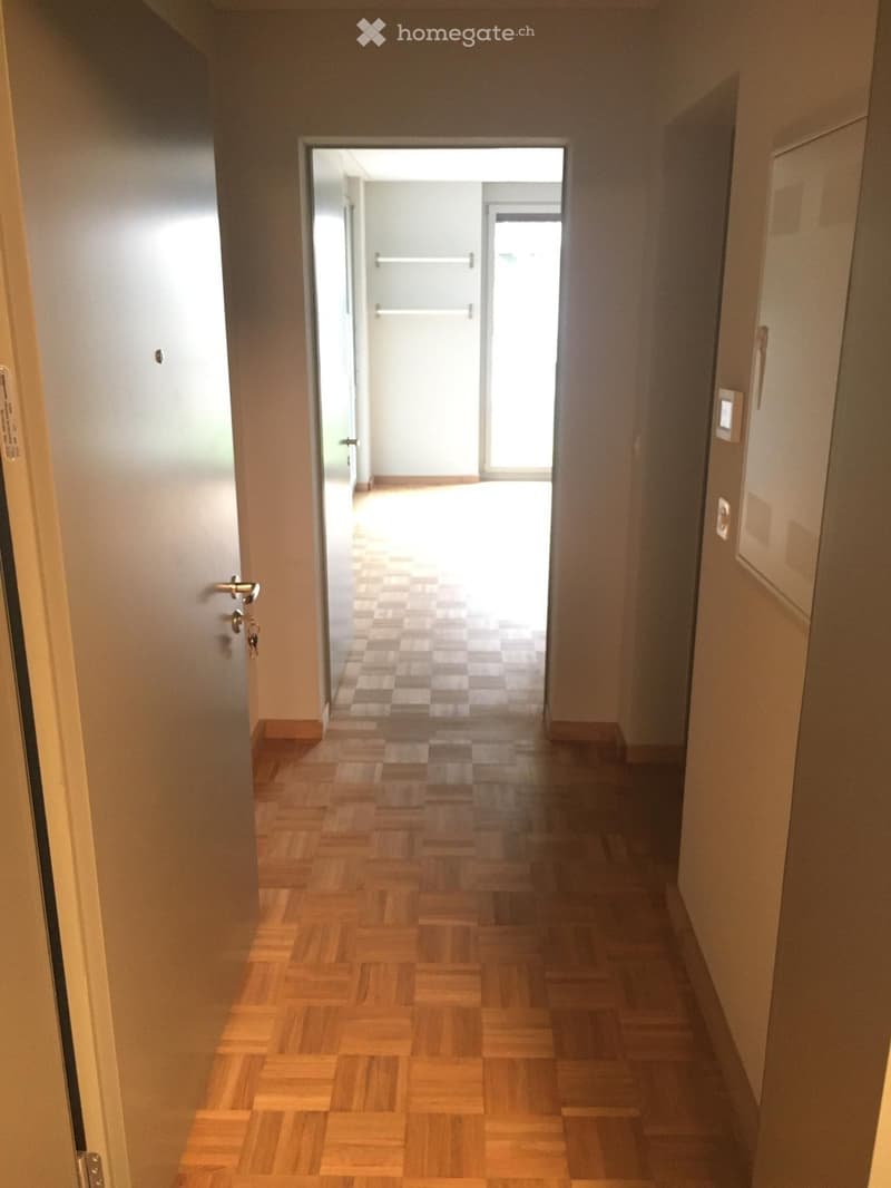 A narrow hallway with wooden flooring and light-colored walls, leading to an open area with a tiled floor and a door to an outdoor space.