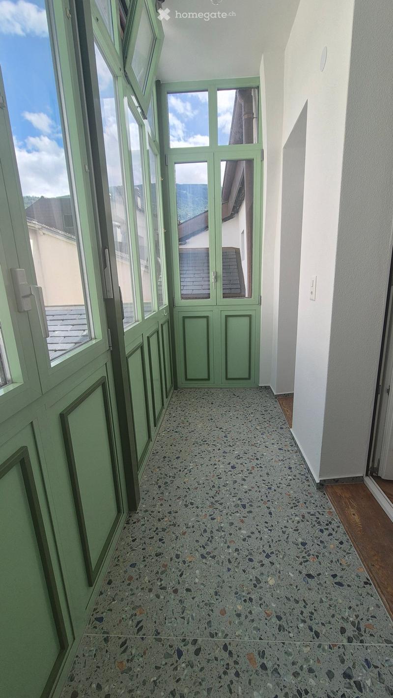 Bright hallway with green double doors leading outside, large windows on one side, and a colorful mosaic tile floor.