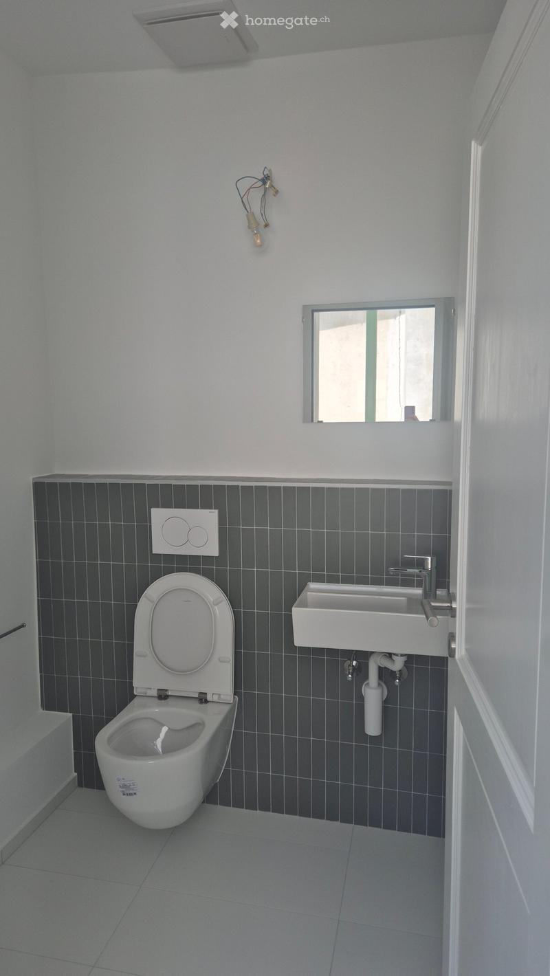 Modern bathroom with white toilet, white sink, and dark grey tiled walls. Features a small window above the sink and a light fixture on the wall.