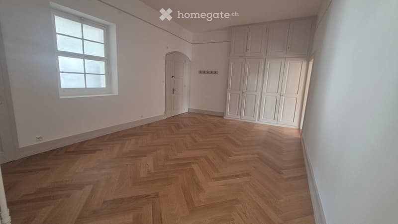 Empty room with wooden herringbone flooring, white walls, large window, white built-in closet, and a coat rack on the wall.