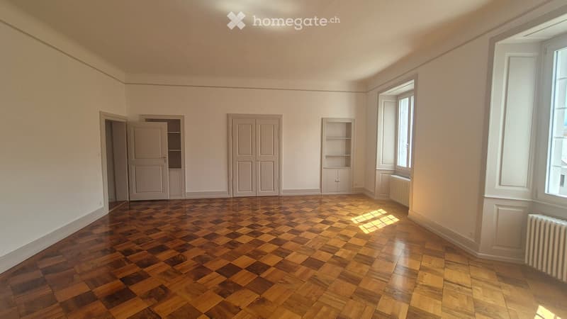 Empty room with wooden parquet flooring, white walls, and large windows allowing natural light. Features include built-in cabinets and a radiator.