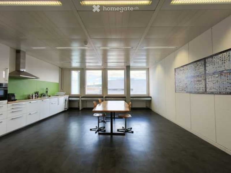 Bright, modern break room with a central table and chairs, kitchen area with cabinets, sink, and microwave, large windows providing natural light, and a colorful artwork on the wall.