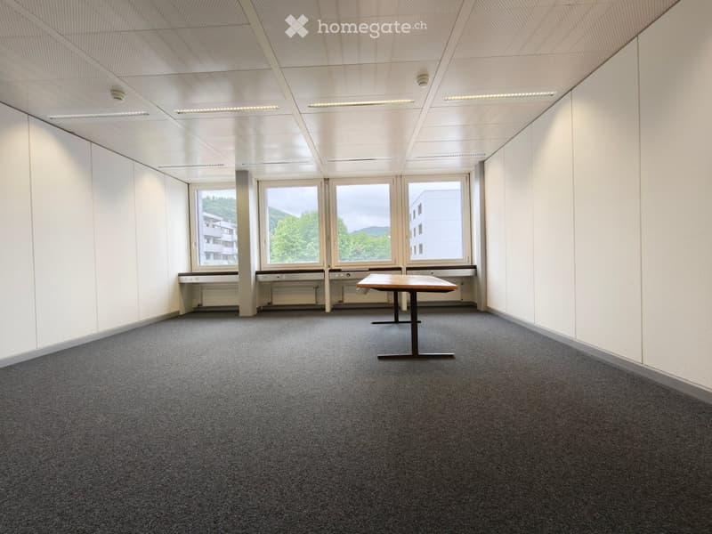 A modern, unfurnished room with grey carpeting, white walls, and large windows offering a view of the outside. A single table is placed in the center.