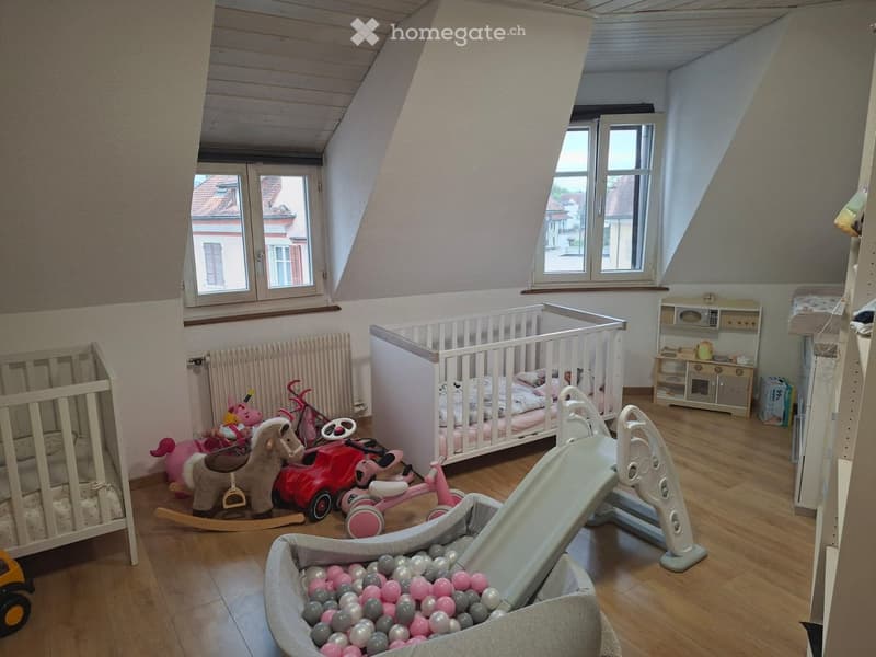 A well-lit attic bedroom with wooden flooring, two cribs, a variety of children's toys including a toy horse, toy car, toy bicycle, and a ball pit, along with a radiator and a small cabinet.